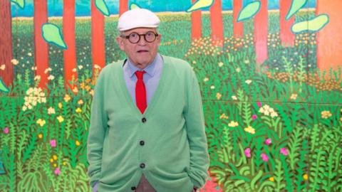 Hockney in a green cardigan, blue shirt, and orange tie in front of one of his paintings 