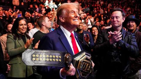 Donald Trump grins while holding a UFC belt handed to him by UFC fighter Jon Jones. He is surrounded by a crowd including Elon Musk and Tulsi Gabbard. 