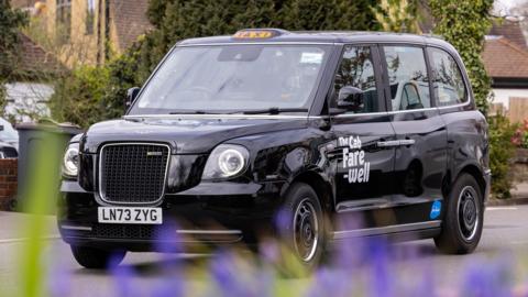 A black London taxi drives down a road. Written on the side is "The Cab fare-well"