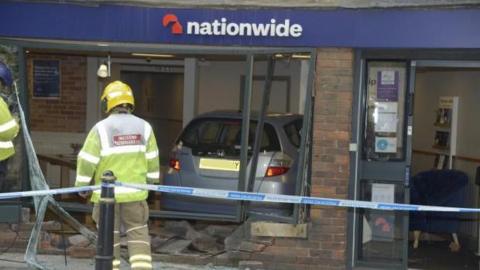 A firefighter stands in front of Nationwide, behind a blue and white striped cordon. The window of the bank is completely smashed, with bricks and glass strewn about. A silver Honda Jazz is visible inside the branch.