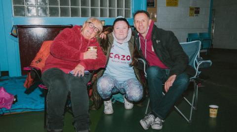 Three fans sit together at a previous sleep out. They have coffee cups and are wearing fleeces and two of them have camping chairs. There is a sleeping bag in the background.