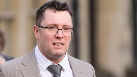 Christopher Trybus. He has short dark hair and is wearing reading glasses, a white collared shirt, a grey tie and a grey suit jacket.