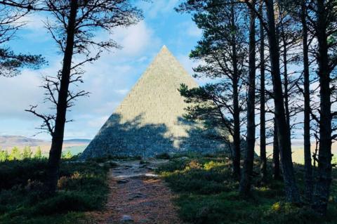 Pyramid-shape cairn at the edge of woodland, with the shadow of trees across the structure.