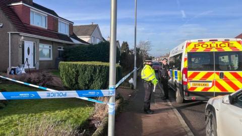 Police tape runs between a house and the road signs outside the property. A police officers stand guard outside the property by the tape. A police van is parked outside.