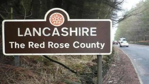A Lancashire road sign. It is brown with white letters saying Lancashire, The Red Rose County. There is a road to the left of it with cars on it.
