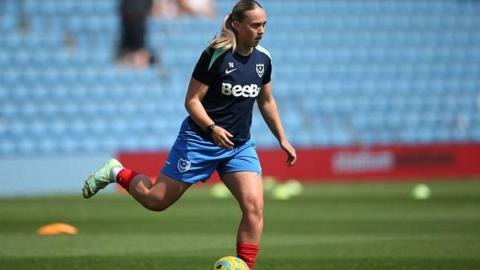 Ella Humphrey playing a game at Portsmouth FC Women. She is on the football pitch running and about to kick a ball. In the background are empty seats.