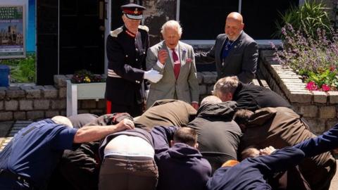 King Charles has arm held by uniformed officer as men scramble in a scrum in front of him