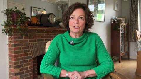 Professor Julia Waters is sitting in her living room looking at the camera, has brown short hair, hazel eyes and is wearing a green jumper.