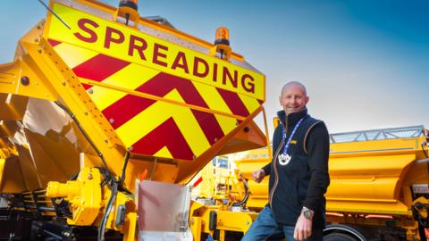 A man stood next to a yellow gritting lorry with the word "spreading" in red capital letter on the back. He is looking at the camera a smiling with a medal round his neck.