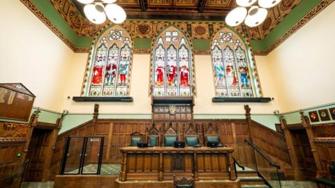 An old, grand looking room with a wooden bench and green leather chairs behind it. Large, decorative stained glass windows are on the wall behind. 
