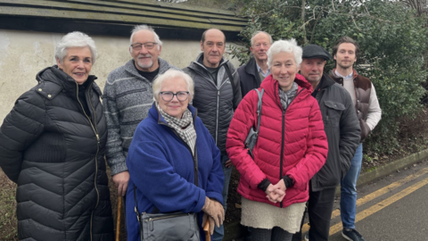 Eight people standing in a group looking into the camera on a road in front of a mud wall and a row of hedges. All are wearing coats or jumpers.