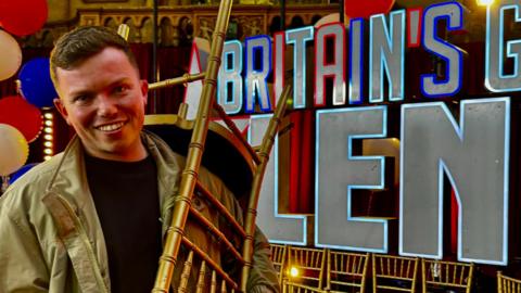 Sonny Green smiling at the camera holding a golden chair over his shoulder with the Britain's Got Talent logo and words in the background. He is wearing a khaki or golden-coloured jacket. He has short brown hair.