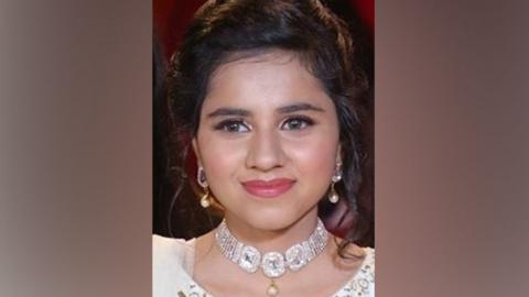 A close up photo of Harshita Brella wearing a white dress, a diamond looking necklace and gold earrings and red lipstick. She is smiling without teeth