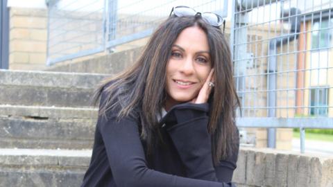 Kathryn Croft, a woman with long dark hair, wears a black jacket and poses on a set of concrete steps in front of a metal railing, her glasses pushed up on the top of her head, as she smiles into the camera.