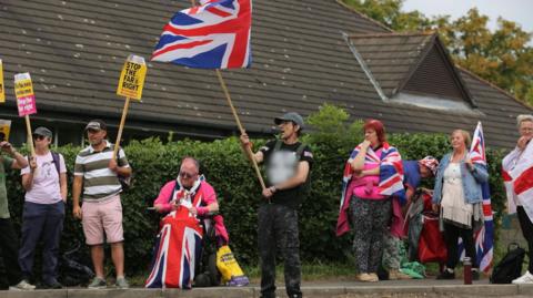 Protestors waving flags and counter protesters with signs saying 'Stop the far right' gather outside a hedge with a hotel behind it