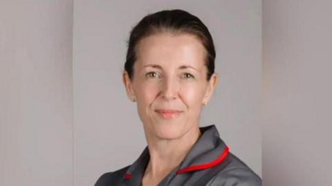 A woman in a grey nursing uniform smiling at the camera. She has dark brown hair pulled back and wears grey studs in her ears. there is red piping around the uniform's collar.