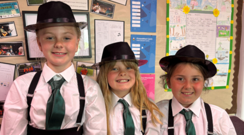 Three young girls, all wearing white shirts, black braces, green ties and black trilby-style hats. The girls on the left and in the centre have blonde hair and the other has dark hair. All of them are smiling at the camera.