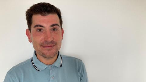 A man with short dark hair looks into the camera. He has light dark stubble on his chin and a light blue top on, in front of a white walll