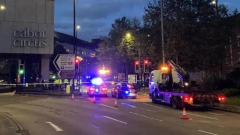 Cabot Circus shopping centre at night. There are several police cars parked next to it with their flashing lights on, and a police cordon is in place at the scene of the crash.
