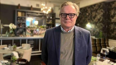 A man with slicked back blond, greying, hair and black-rimmed glasses looks straight down the camera. He's wearing a navy jacket over a grey jumper and white dress shirt. He's standing in a restaurant - behind him are tables and chairs with small green plants. 