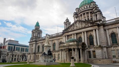 A stock image showing the exterior of Belfast City Hall during the day. It is an ornate grey building with copper green tower domes.