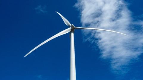 A wind turbine viewed from below, looking up at three blades from the ground. It is mostly a clear day with cloud seen in the blue sky above.