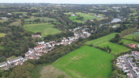 An aerial view of fields, trees and two separate rows of houses. The sea can be seen further afield.