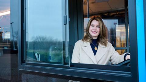 Kim McGuinness sitting behind the steering wheel of a bus. She is wearing a white coat and a blue top. She has one hand on the steering wheel and is looking out of the window and smiling at the camera.