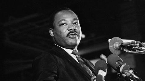 A black and white photograph of the late Dr Martin Luther King Jr. He is standing at a podium, surrounded by microphones, as he speaks, looking to the right of the stage.