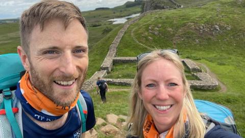 Luke Hames-Brown has a mousey brown beard and short hair and Kate has blonde, shoulder length hair. They're both carrying rucksacks and MND Association snoods 