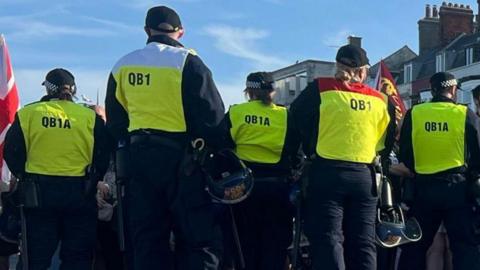 Rear view of a line of male and female police officers wearing hi-vis vests and holding riot helmets. A union flag and a Royal standard flag can be seen above their heads, held by people on the other side of the line