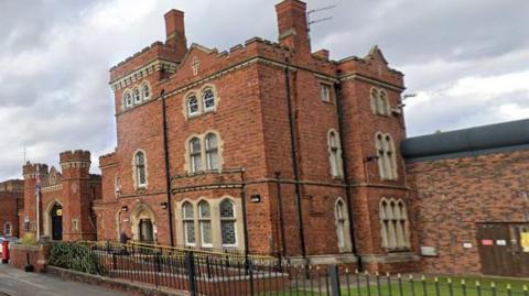 Victorian frontage of the red-bricked HMP Lincoln prison building with black railing in the foreground and high wall to the side