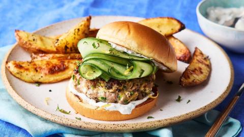 A burger filled with lamb, feta sauce and sliced cucumber, on a plate with golden wedges