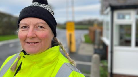 Toll keeper Nikki Wilcock with blond hair in plaits and brown eyes in a hi-vis jacket and black and white woolly bobble hat 