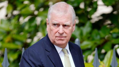 A close-up image of Andrew Mountbatten-Windsor. He is an older man with short white hair. He is wearing a dark blue suit jacket, a white shirt and a pale green and yellow patterned tie. He is looking away from the camera with a neutral expression on his face.