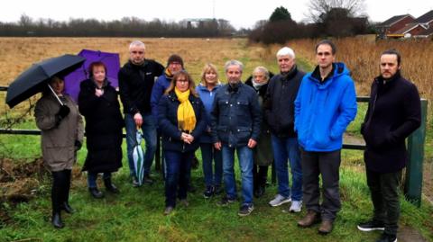 A group of residents standing near the site of the proposed park and ride. The picture shows six men and five women in front of a gate into a field. Two of the women have umbrellas open. 