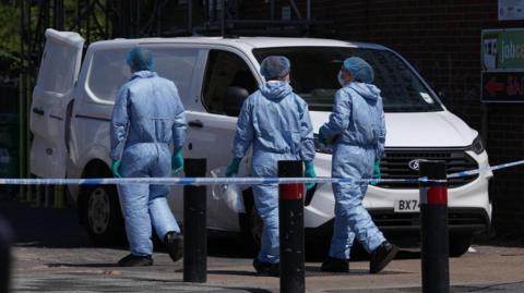 Three people wearing forensic protective suits, hair coverings and gloves walk beside a white van behind a police cordon. Bollards and tape are visible in the foreground, with a building and fencing in the background.
