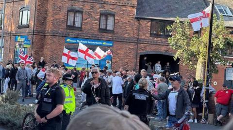 Protesters are gathered in a Worcester street with a brick building behind them. They are standing on the pavement with St George's and union jack flags. Police are also at the scene, and the officers are standing at the edge of the group.