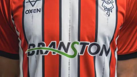 Photo of a man's chest wearing Lincoln City Football Club's latest shirt - red and white stripes with a black trim - which features a black line of binary numbers spelling out 'We Are Imps'. 