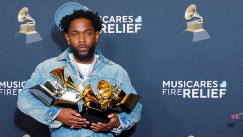 Kendrick Lamar wearing a denim jacket and light denim cap holds four grammys in his hands