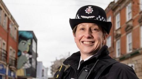 Rachel Nolan smiles at the camera. It is a head-and-shoulders shot. She is wearing her uniform and police hat and is standing in a high street. The street is out of focus.