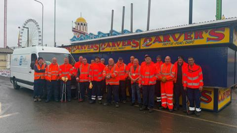 Part of the cleaning team standing in front of a chip stall. They are wearing orange high visibility jackets and some are carrying litter picking tools