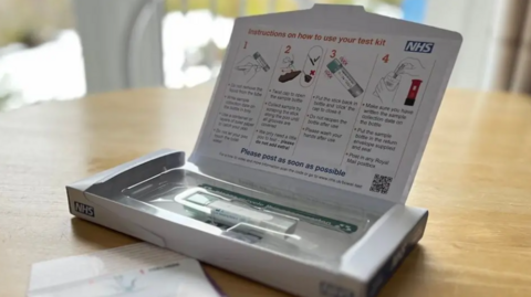 A close-up of a home-testing kit, consisting of a small white cardboard box containing a stick and a container in packaging, laid open on a dining room table