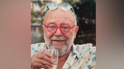 A man with short grey hair and a trimmed beard is seated outdoors, holding a glass of a light-coloured sparkling drink. The person is wearing bright red round glasses, with a second pair of turquoise glasses resting on top of the head. The clothing is a light shirt with a colourful floral pattern. The background is softly blurred, showing greenery and hints of buildings, suggesting a garden or outdoor social setting.