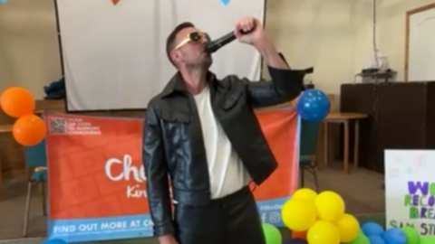 Sam Stephens holding his microphone aloft. He has short black hair, is wearing a black leather jacket, black glasses and is standing in front of a projector screen.