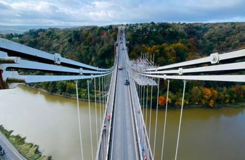 An aerial view of the Clifton Suspension Bridge taken from one of the towers, showing Leigh Woods in the distance and the River Avon below