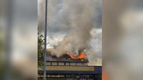 The roof of a building is burning, with flames and smoke seen rising into the sky. 