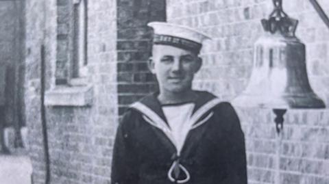 An archive black and white image of Leading Seaman Jack Mantle, dressed in his navy uniform. He is standing next to a bell. He is smiling.