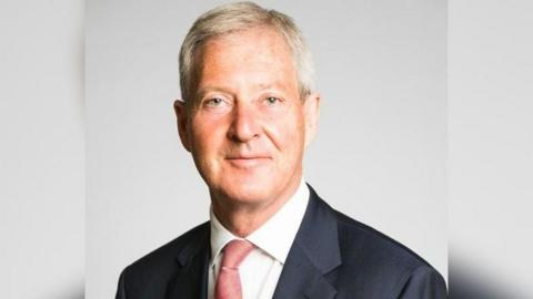 A headshot of Tim Oliver in which he is looking at the camera. He is wearing a dark suit, white shirt and red tie and is smiling.