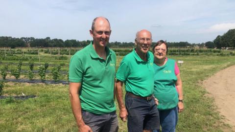 Peter is standing in the centre, with his wife to the left of him and his son to the right.
They are all wearing green tops and in the background is their pick your own farm.
The sky is a mix of blue and wispy thin cloud.